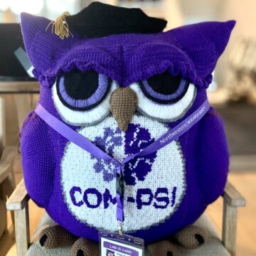 A purple owl with the COM-PSI logo on its belly, wearing a graduation cap and lab name tag.